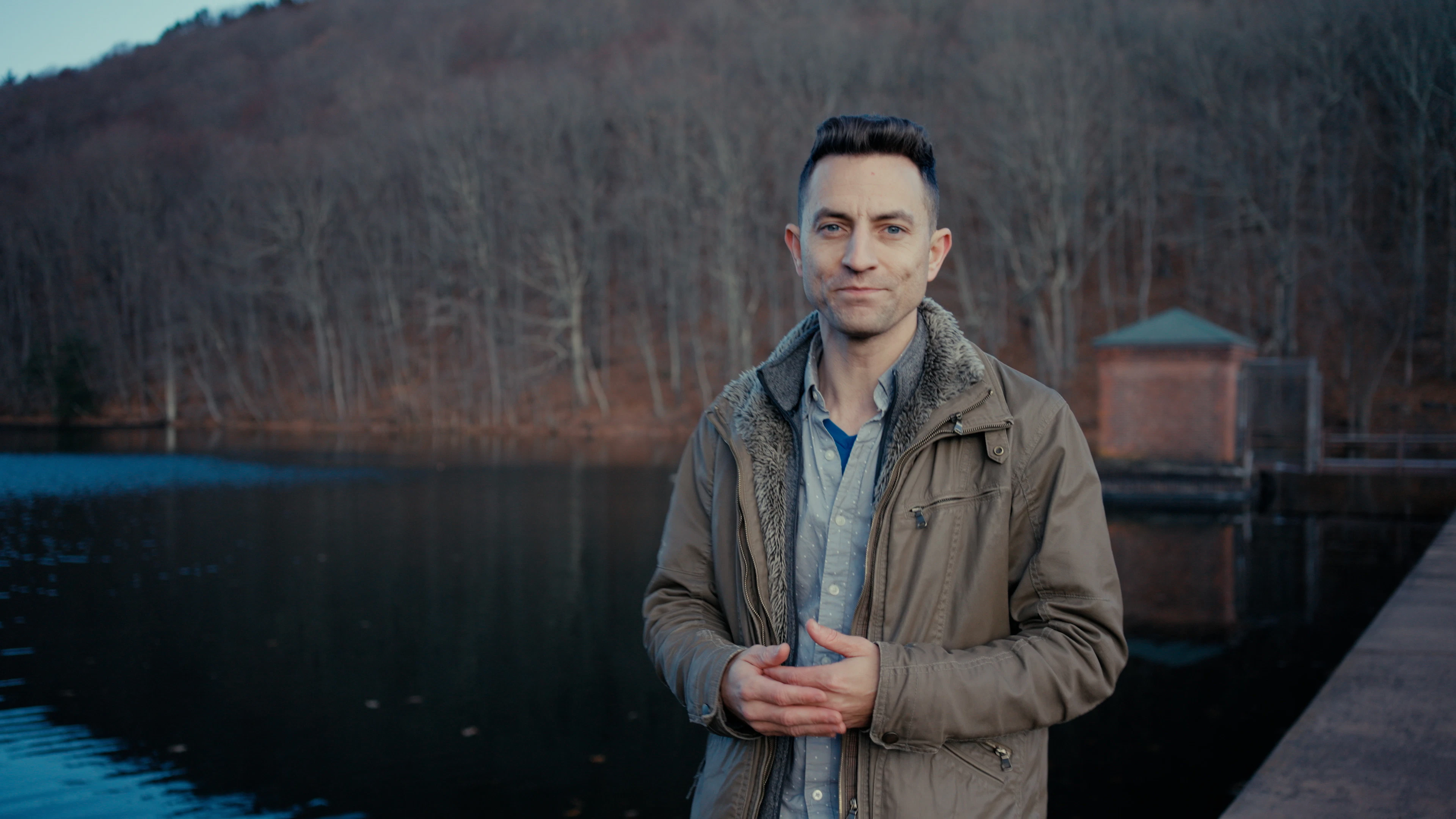 The winner of the Artlist 2024 $100K Fund competition stands by a tranquil lake with bare winter trees and a small brick structure in the background, an inspiring scene for content creators and video editing enthusiasts.